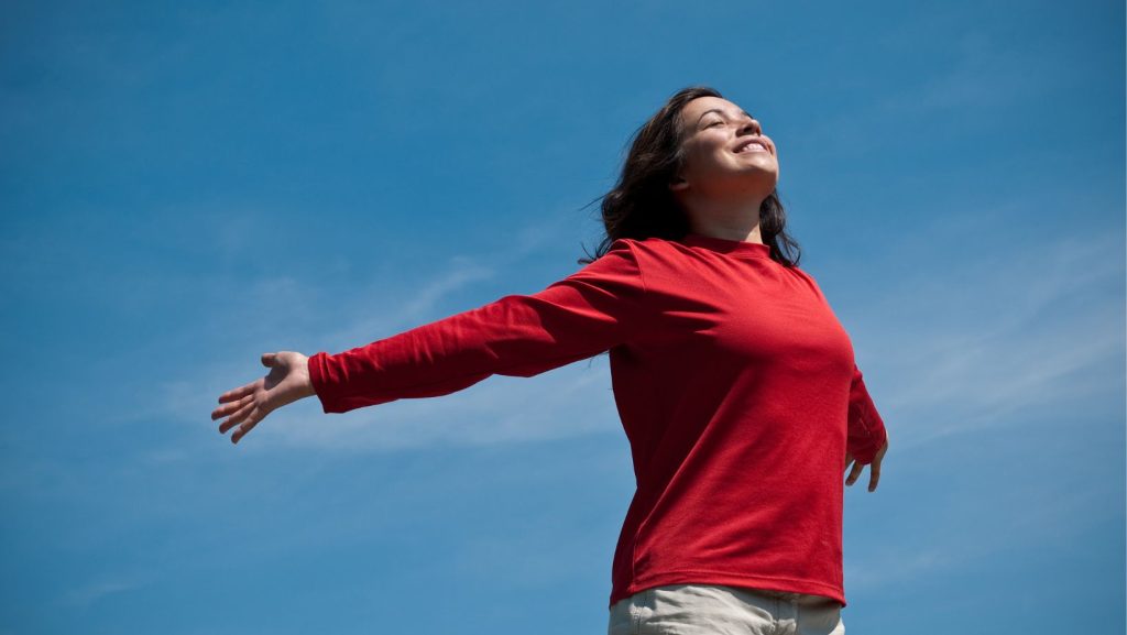 A woman in a red top with arms wide open, embracing the sky, symbolizing freedom and relief.